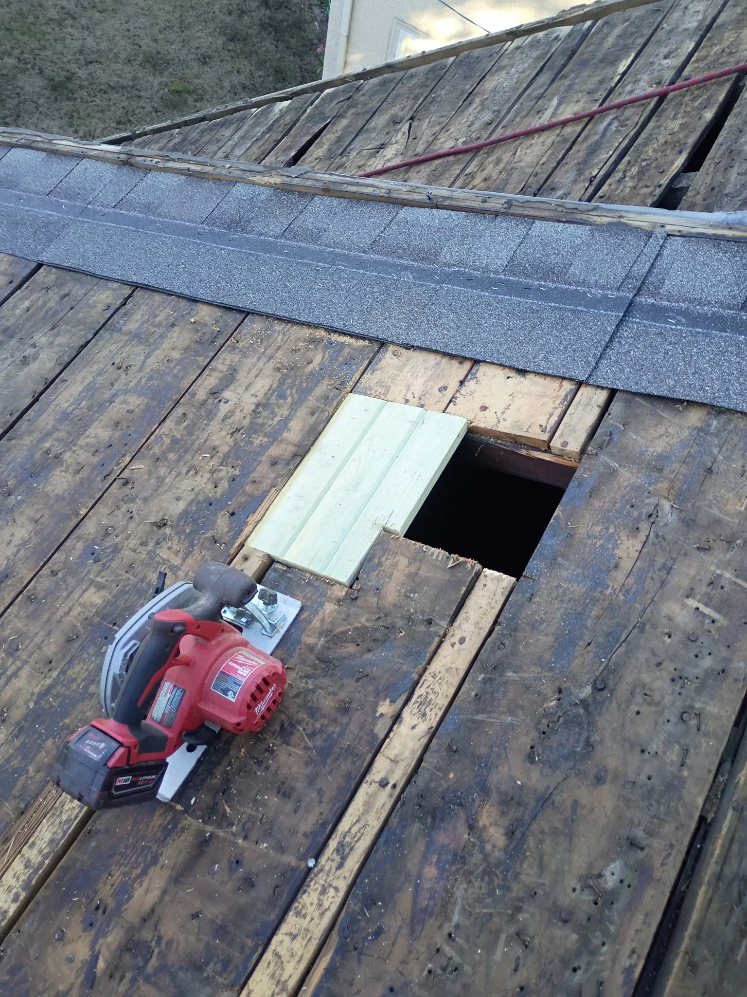 Milwaukee circular saw cutting roof decking with new lumber boards fitted into opening beside gray asphalt shingles