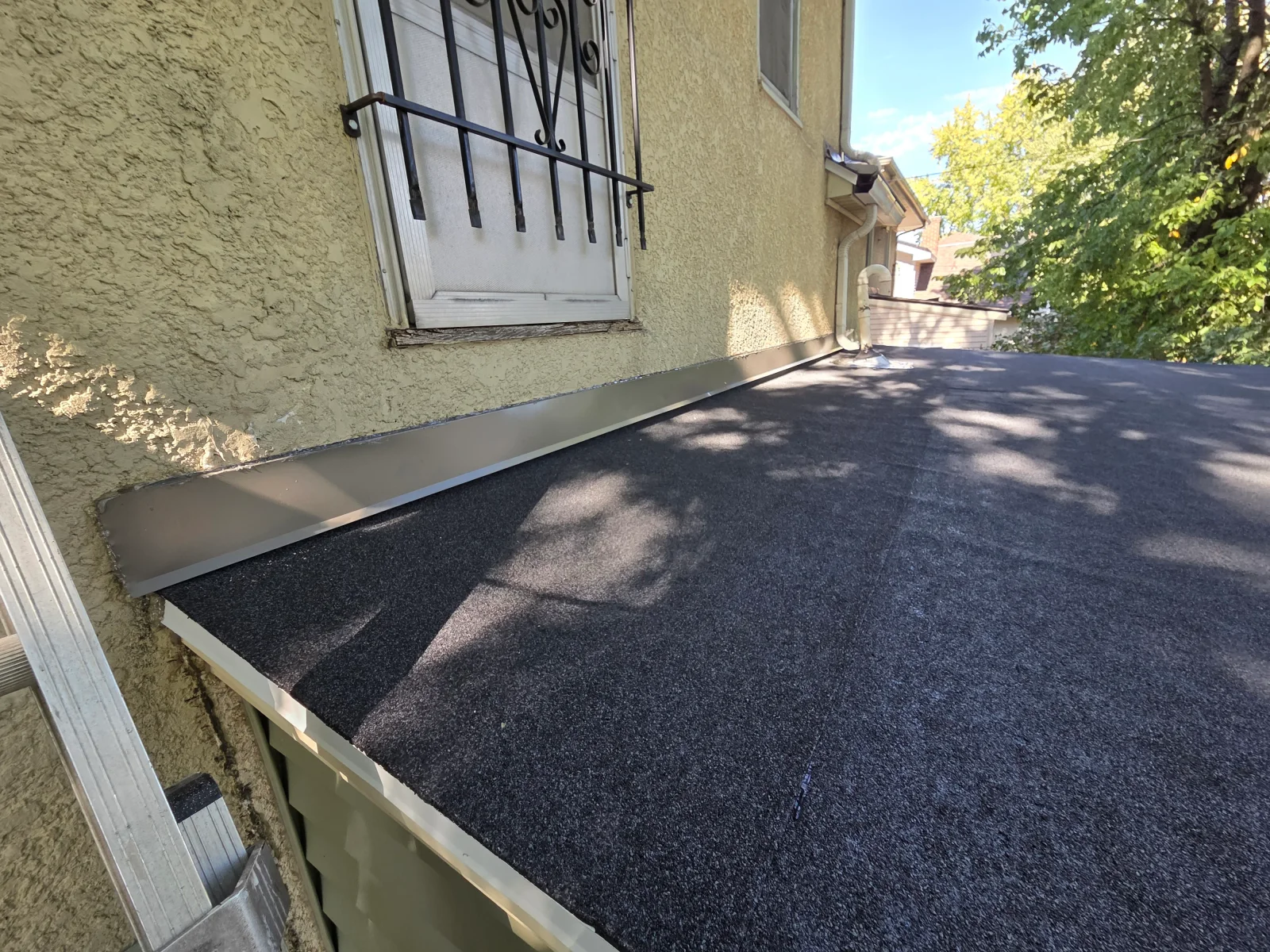 Low-slope roof with dark rolled roofing membrane and metal wall flashing against textured stucco wall