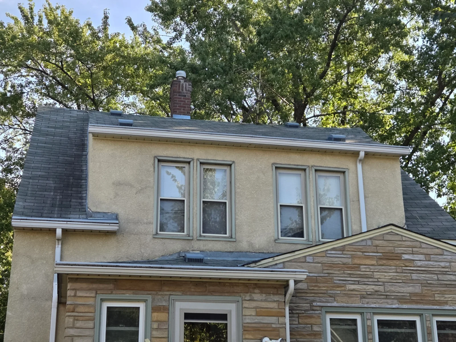 Two-story stucco home with dark asphalt shingle roof, brick chimney, roof vents, and white gutters