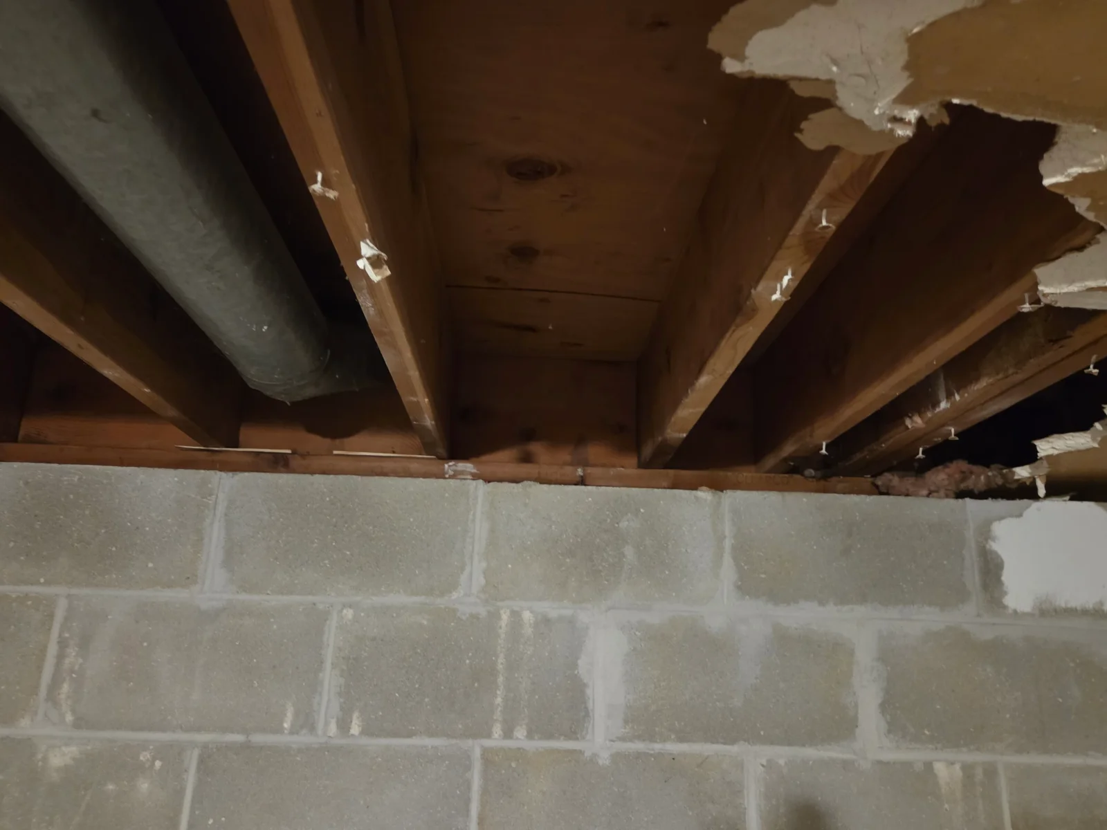 Exposed wood joists and subfloor boards above concrete block foundation wall with metal duct
