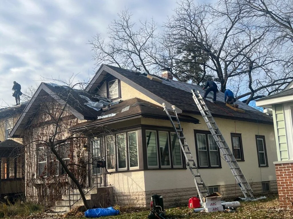 Crew replacing asphalt shingle roof on stucco house with aluminum extension ladders and exposed underlayment