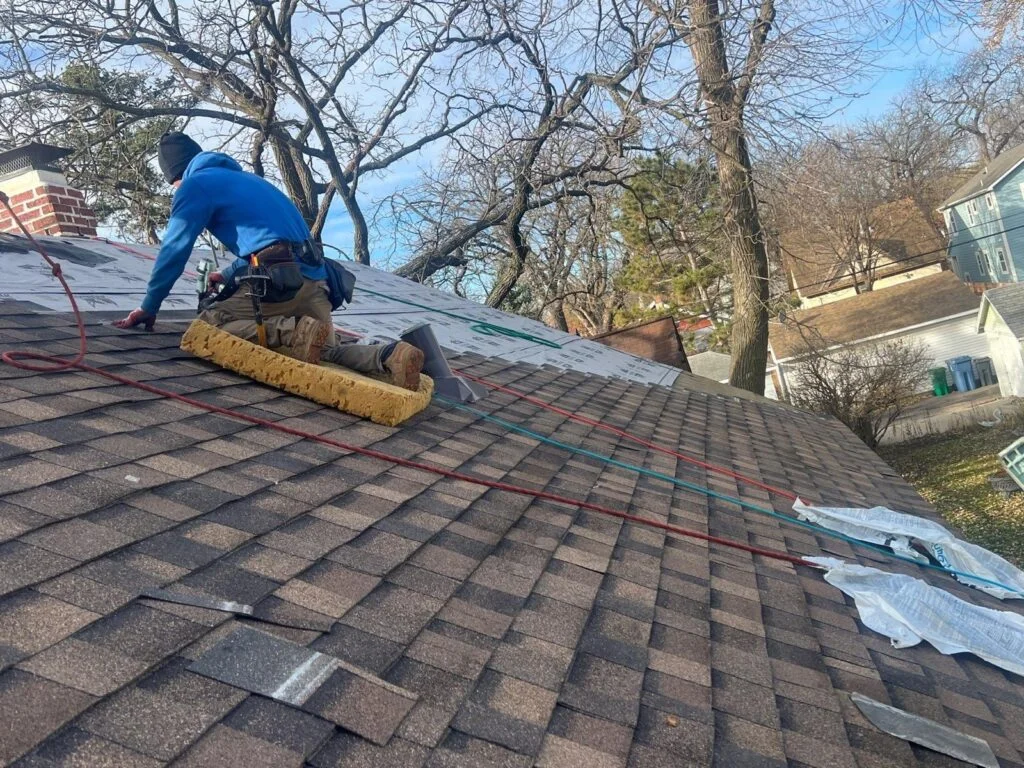 Roofer kneeling on pitched roof installing brown asphalt shingles over white synthetic underlayment with a metal vent flashing