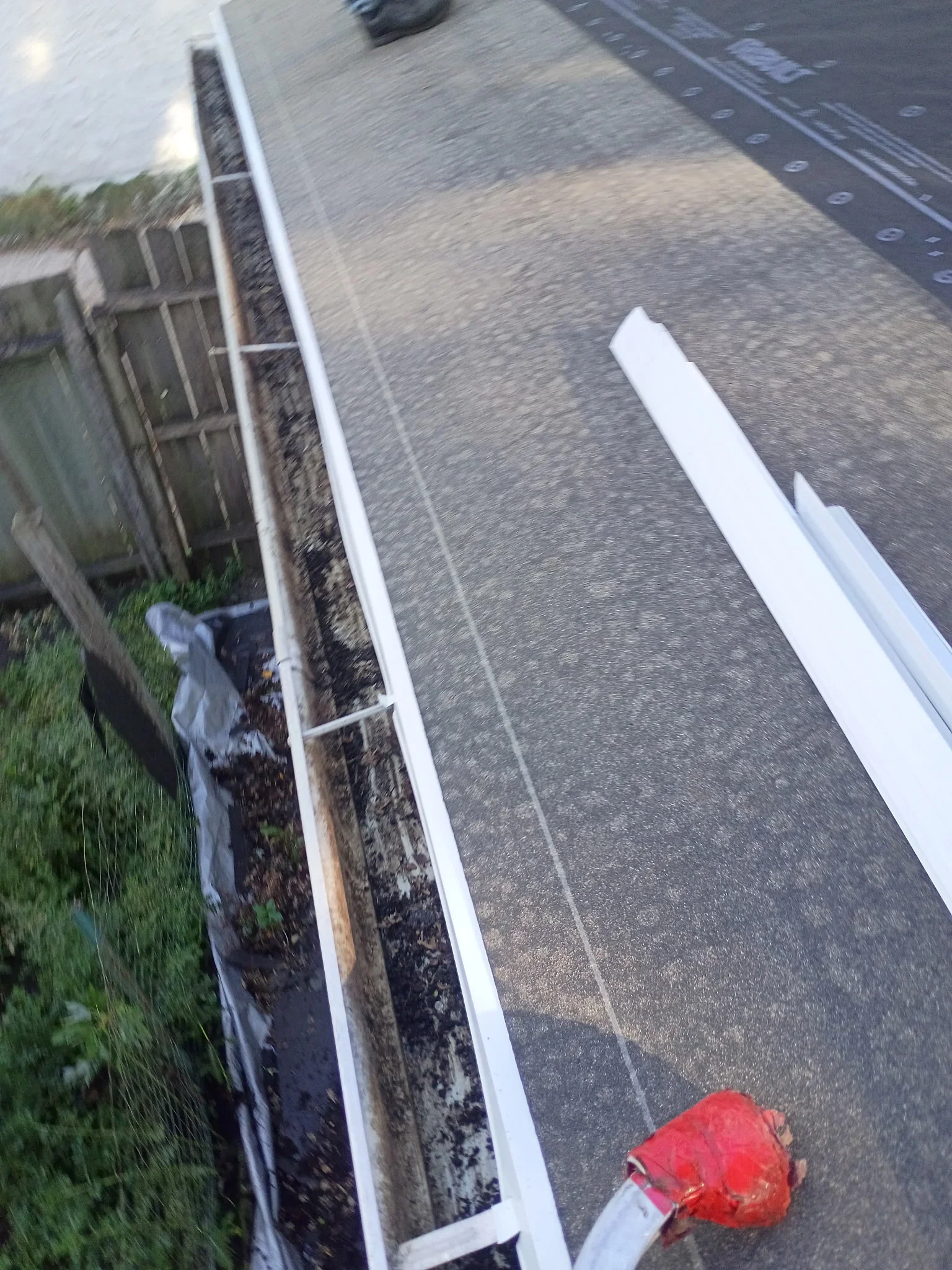 White vinyl gutter packed with dark leaf debris being cleared with a red-handled gutter scoop along an asphalt shingle roof edge