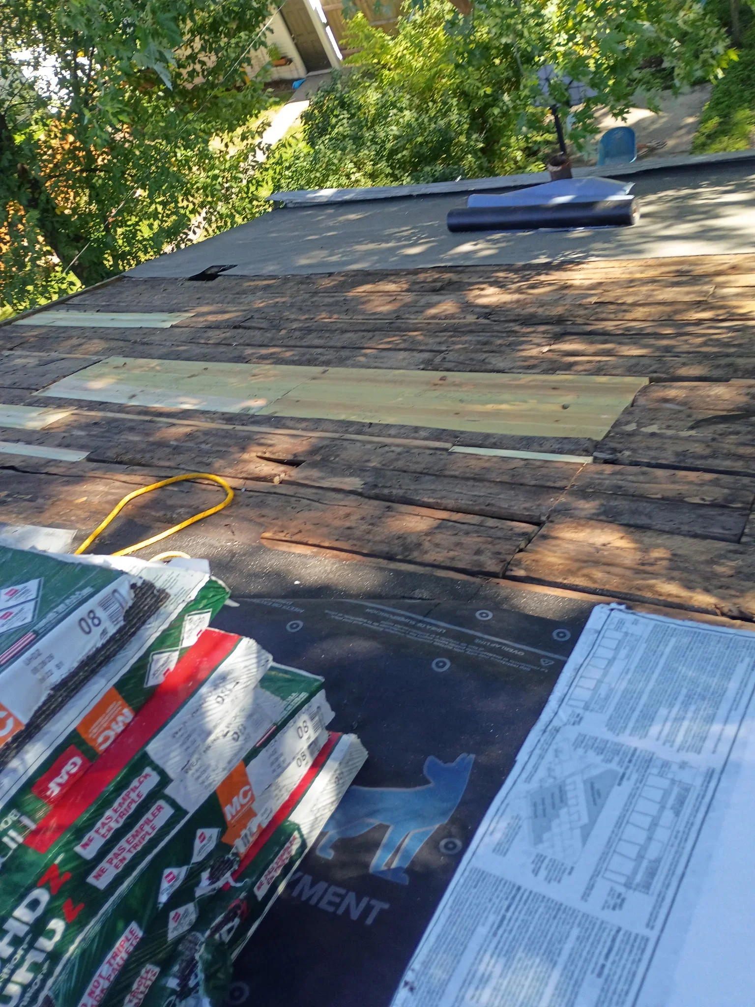 Roof deck with weathered boards, green plywood patches, black synthetic underlayment, and stacked asphalt shingle bundles