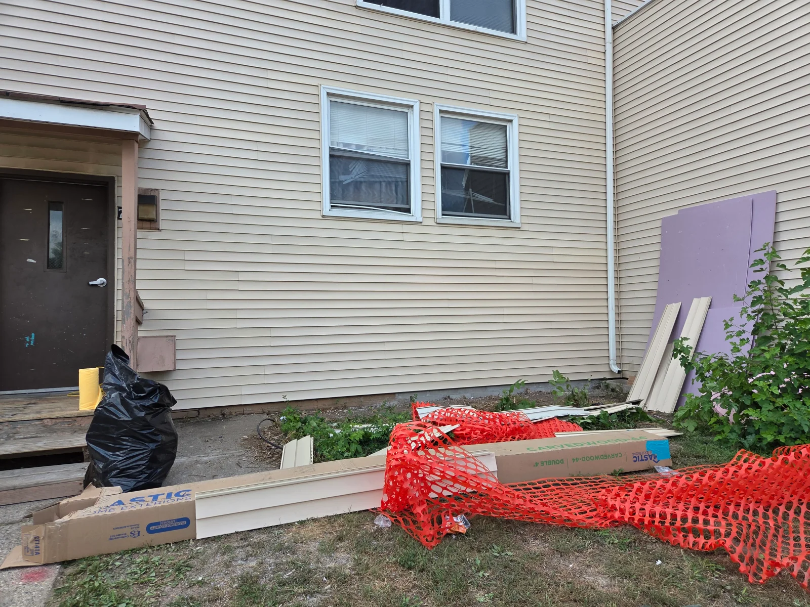 Cream vinyl siding with white window trim and staged vinyl trim pieces in cardboard packaging beside orange safety fencing