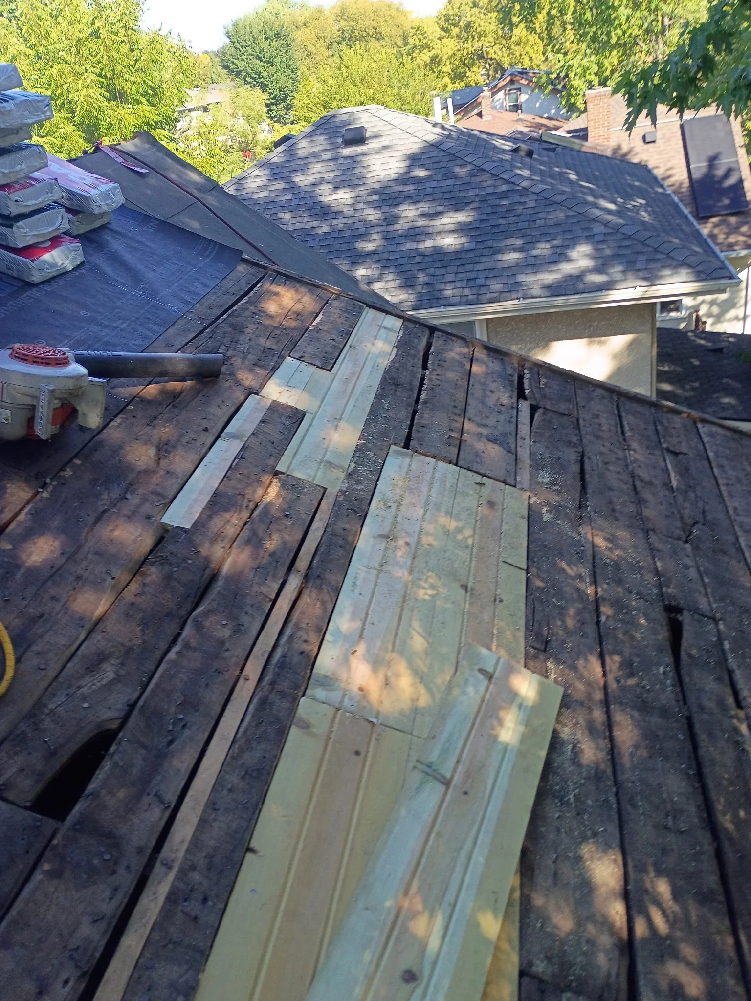 Roof deck with old weathered boards partly replaced by new lumber planks, black underlayment, and pneumatic nailer on site