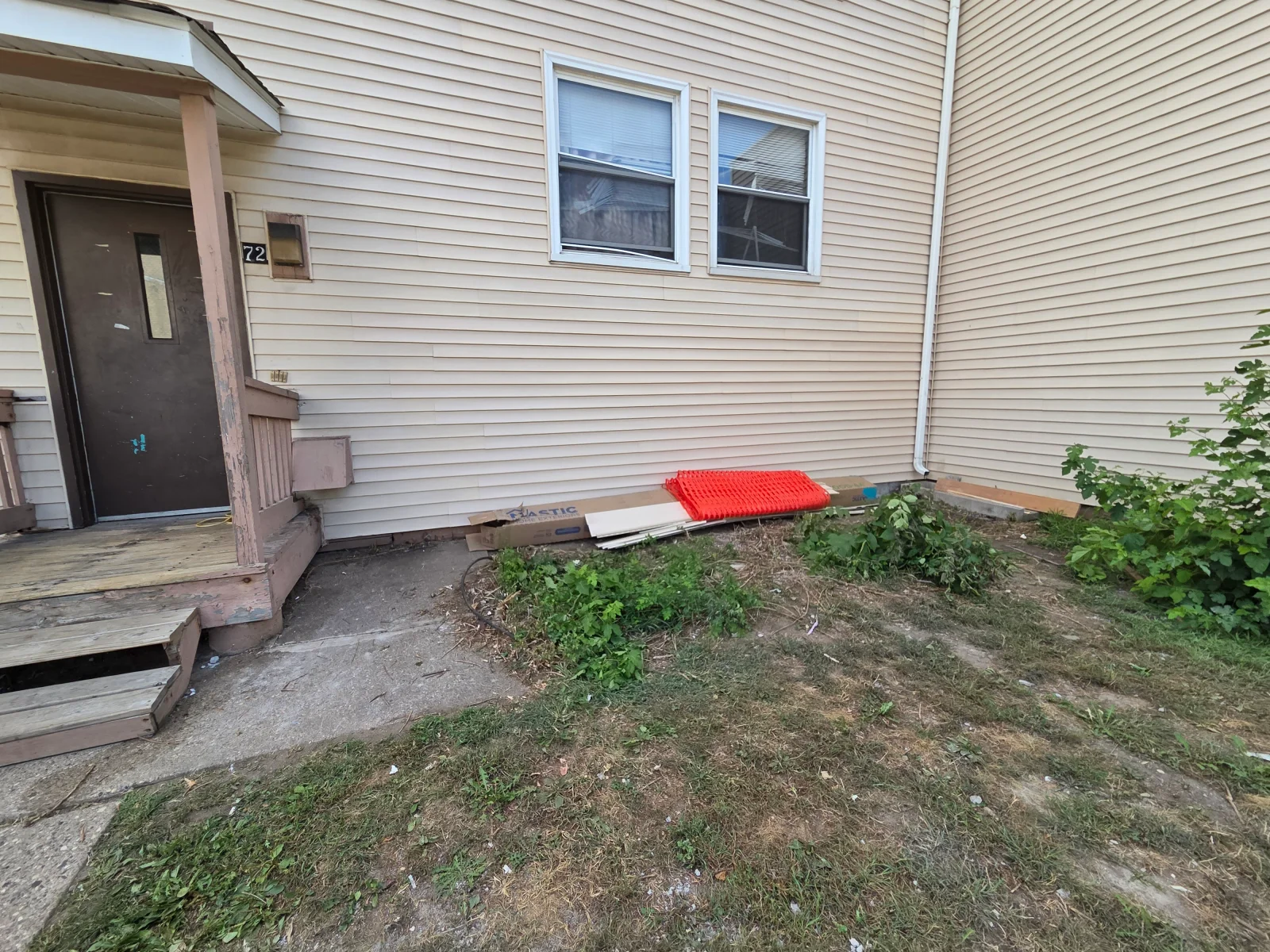 Cream vinyl siding wall with white-trimmed windows, downspout, and debris and orange plastic fencing at the foundation base