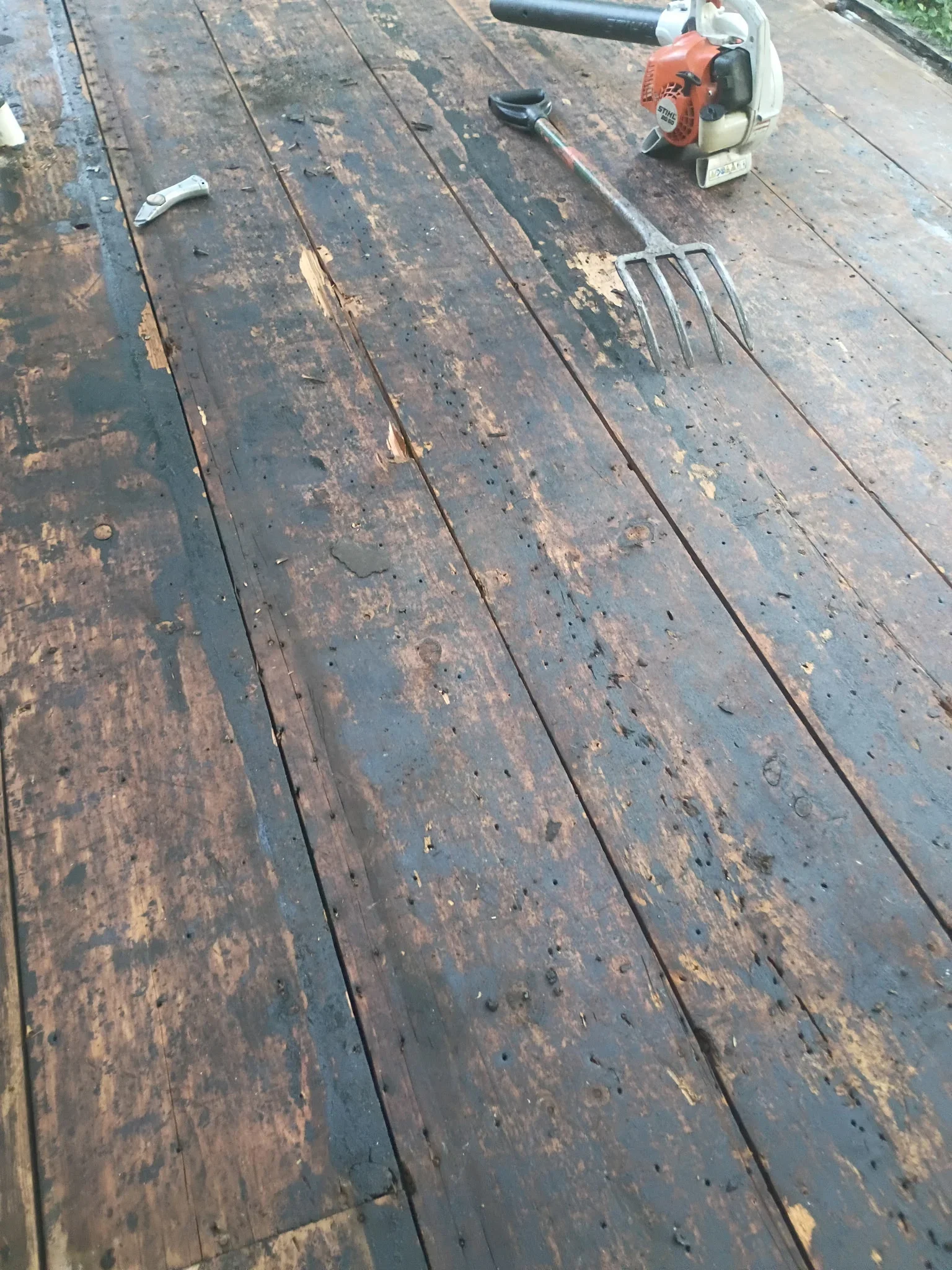 Weathered wood plank surface with dark flaking coating partially stripped, debris and a STIHL blower on the boards
