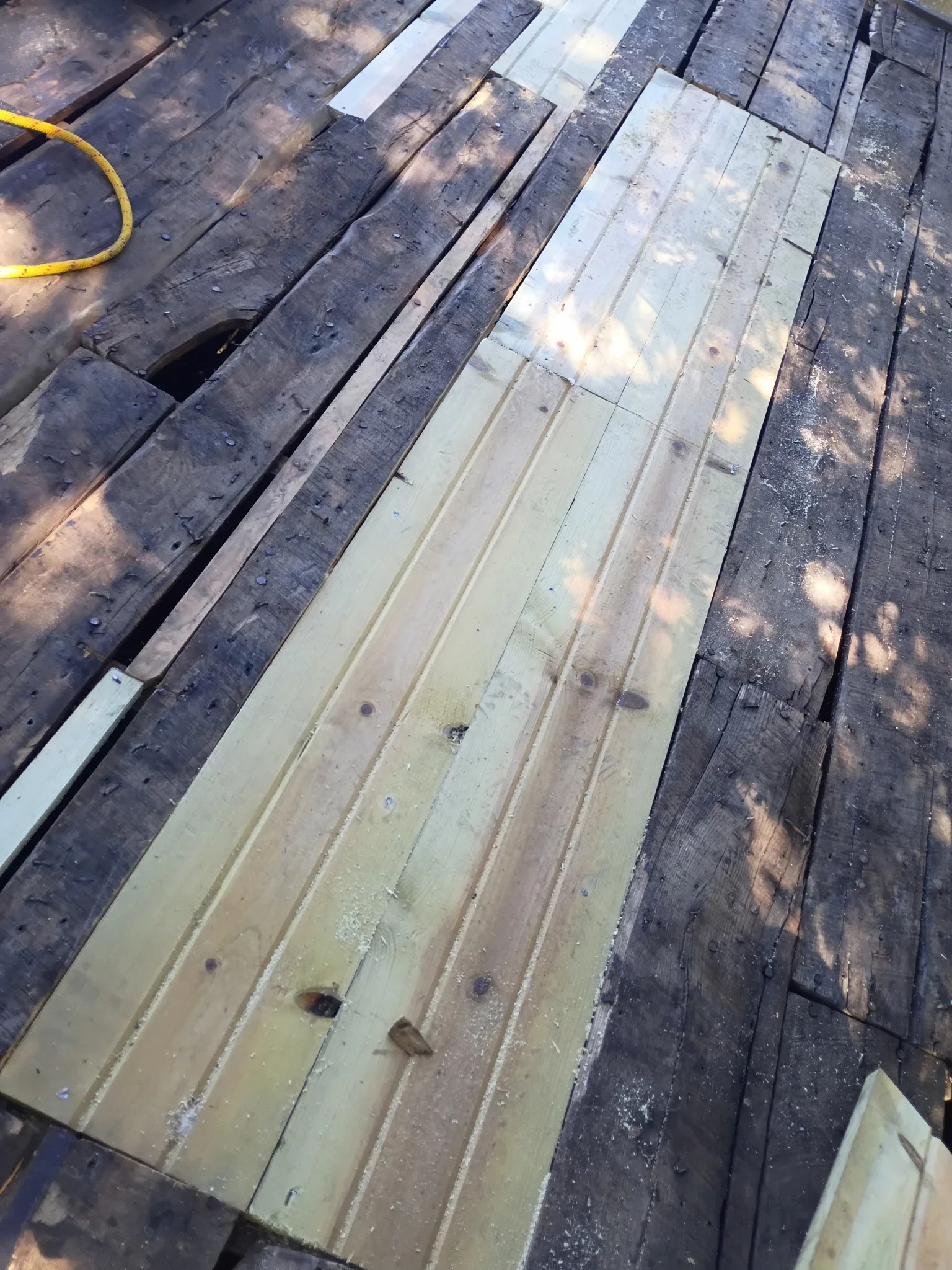 Pressure-treated pine deck boards being installed over weathered gray decking with exposed nail heads and fresh sawdust