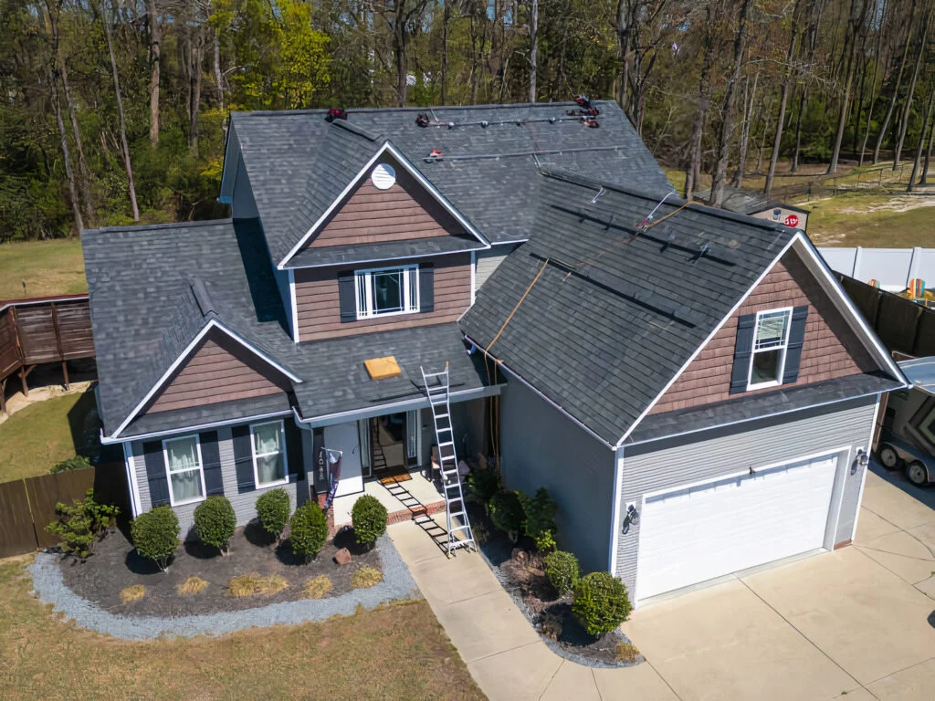 Aerial view of a home with dark gray asphalt shingle roof being installed, brown lap siding, and white trim