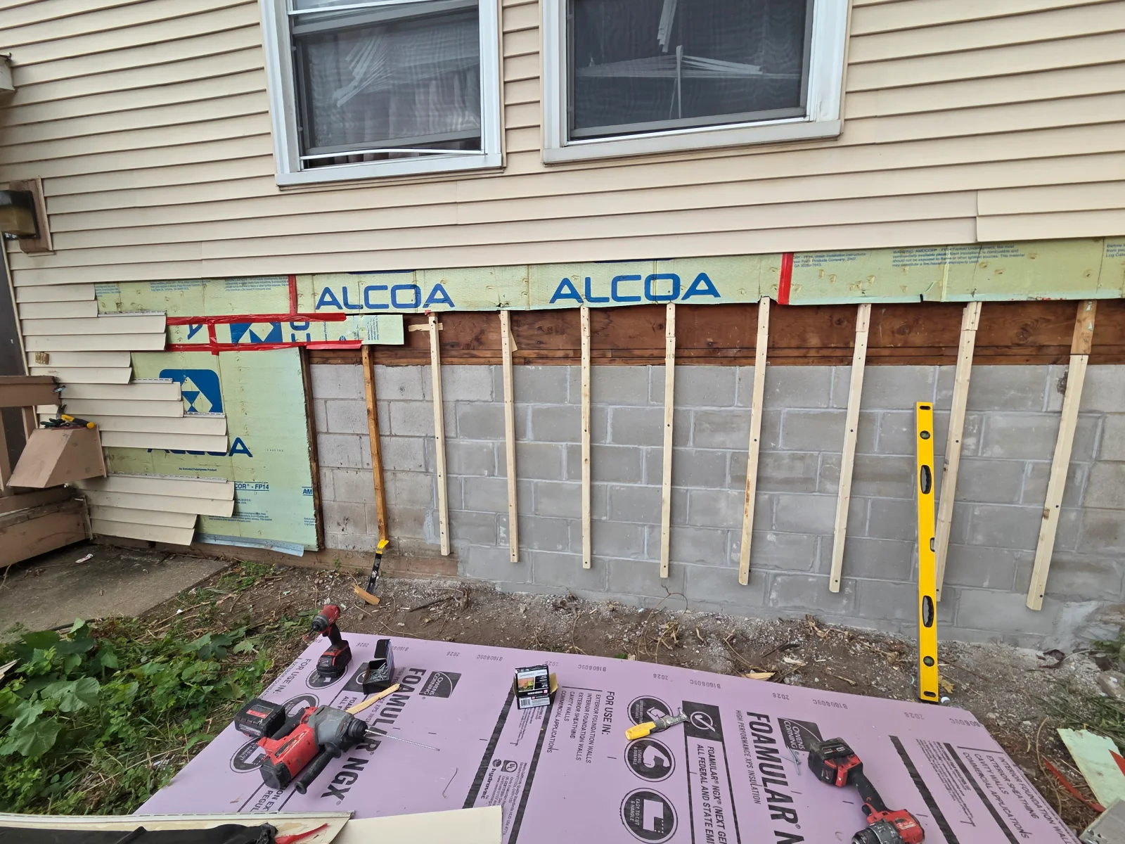 Rigid foam insulation boards and vertical wood furring strips installed on a concrete block foundation wall beneath vinyl siding